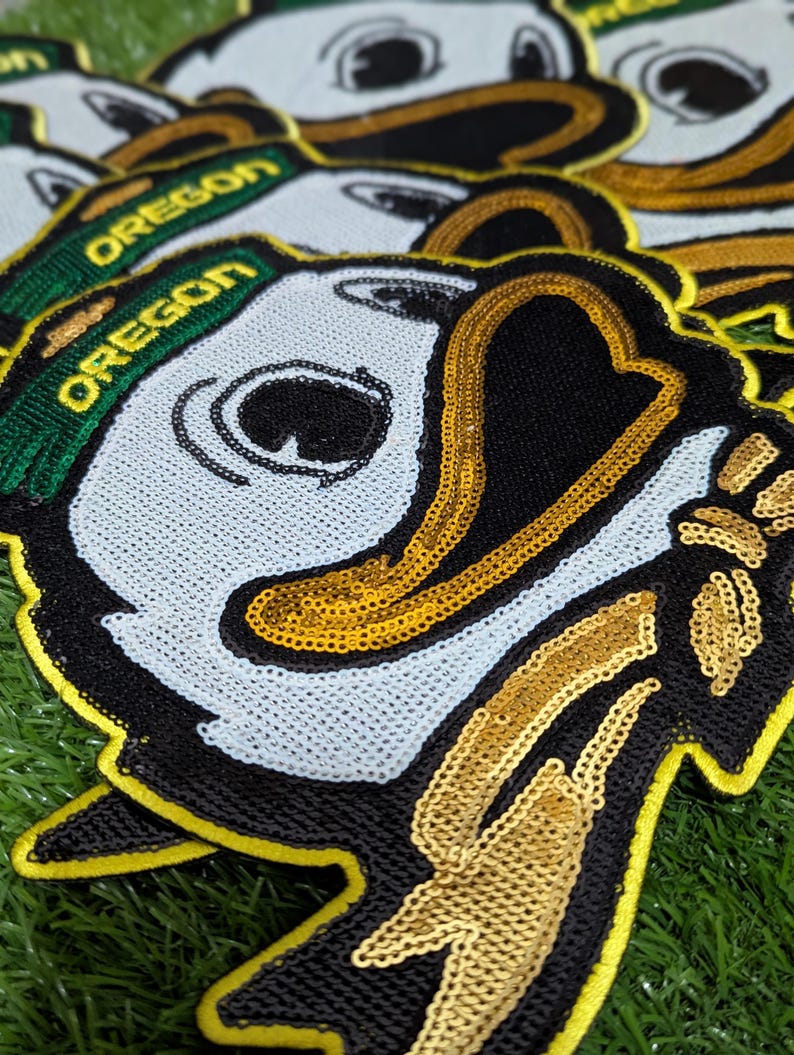 May include: Close-up of several sequined patches depicting the Oregon Ducks mascot. The patches have a white face, black beak, and a green and gold "OREGON" banner. The patches are on a green surface.