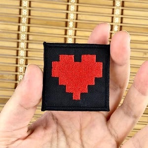 K&ouml;nnte beinhalten: Ein schwarzer quadratischer Aufn&auml;her mit einem pixeligen roten Herzdesign. Der Aufn&auml;her wird in einer Hand gehalten und zeigt das gestickte Herz vor dem schwarzen Hintergrund. Geeignet f&uuml;r Kleidung oder Accessoires.