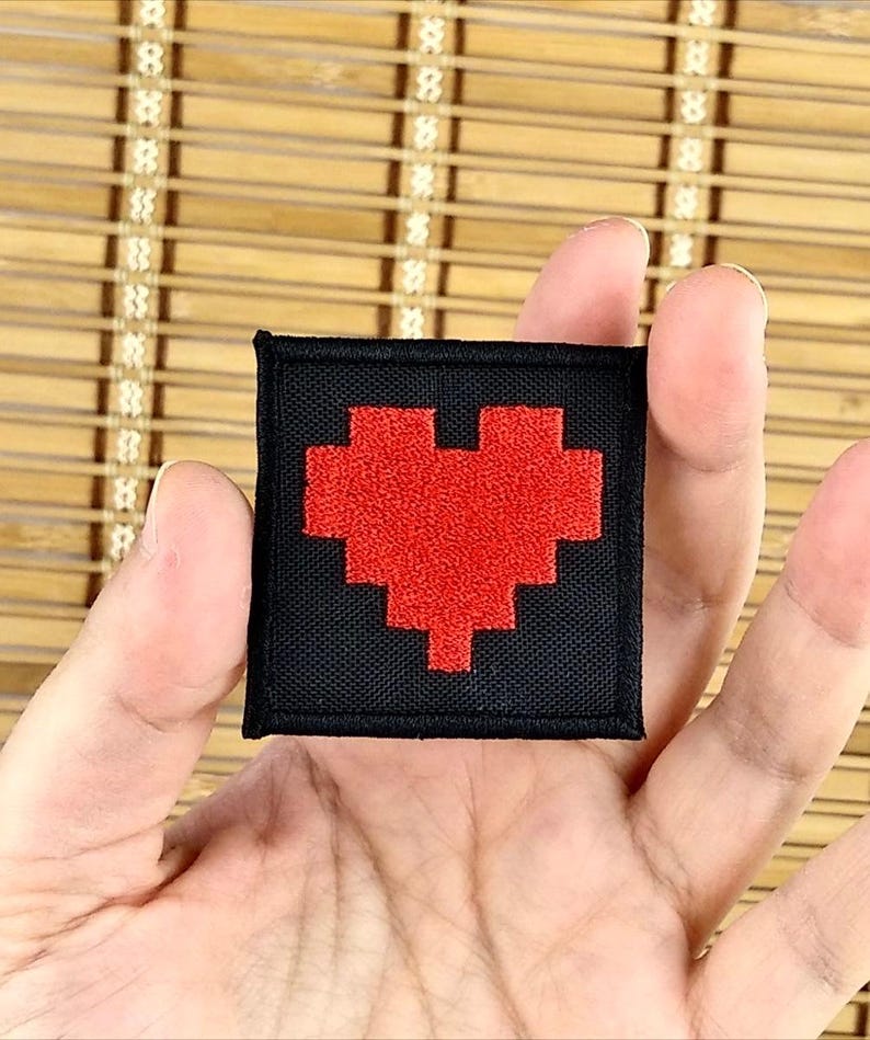 K&ouml;nnte beinhalten: Ein schwarzer quadratischer Aufn&auml;her mit einem pixeligen roten Herzdesign. Der Aufn&auml;her wird in einer Hand gehalten und zeigt das gestickte Herz vor dem schwarzen Hintergrund. Geeignet f&uuml;r Kleidung oder Accessoires.