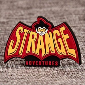 May include: A colorful emblem featuring the words "STRANGE ADVENTURES" in yellow, outlined in black, set against a red bat-wing design. A cartoon character with a hat and glasses is at the top. The emblem is on a textured gray surface.
