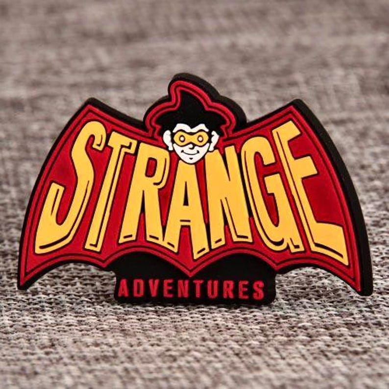 May include: A colorful emblem featuring the words "STRANGE ADVENTURES" in yellow, outlined in black, set against a red bat-wing design. A cartoon character with a hat and glasses is at the top. The emblem is on a textured gray surface.