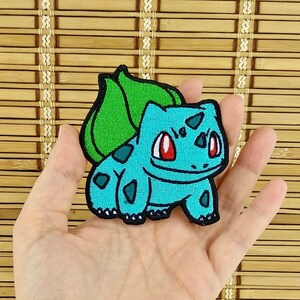 K&ouml;nnte beinhalten: Ein t&uuml;rkisfarbener, gestickter Aufn&auml;her mit einem Bulbasaur-Design. Der Aufn&auml;her hat ein gr&uuml;nes Blatt auf dem R&uuml;cken und rote Augen. Der Aufn&auml;her ist etwa 7,6 cm hoch und 7,6 cm breit.