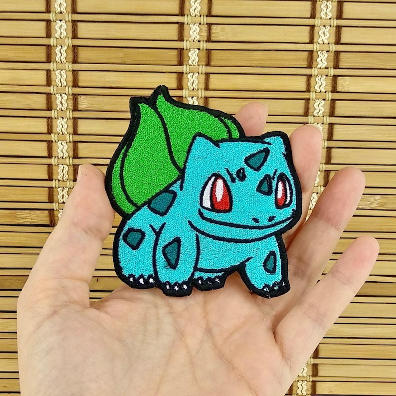 K&ouml;nnte beinhalten: Ein t&uuml;rkisfarbener, gestickter Aufn&auml;her mit einem Bulbasaur-Design. Der Aufn&auml;her hat ein gr&uuml;nes Blatt auf dem R&uuml;cken und rote Augen. Der Aufn&auml;her ist etwa 7,6 cm hoch und 7,6 cm breit.