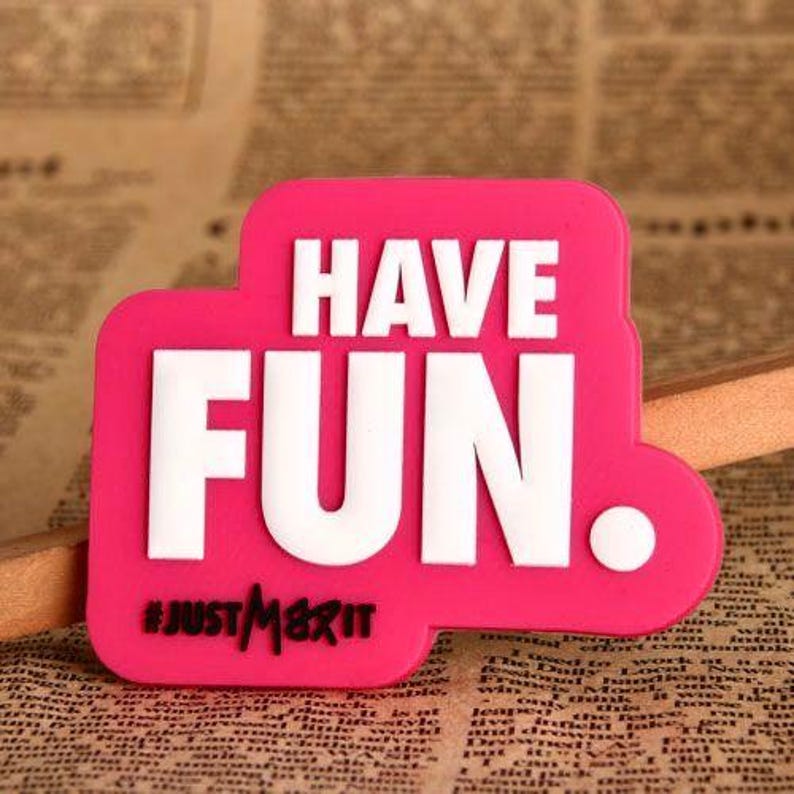 May include: A bright pink rectangular sticker with rounded edges, featuring the words "HAVE FUN" in bold white letters. Below, the hashtag "#JUSTMAXIT" is in black. A white dot is on the right side of the sticker.