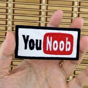 K&ouml;nnte beinhalten: Gestickter Aufn&auml;her mit den Worten "You Noob" in Schwarz und Wei&szlig; auf wei&szlig;em Hintergrund, mit einem roten Rechteck. Der Aufn&auml;her hat einen schwarzen Rand.