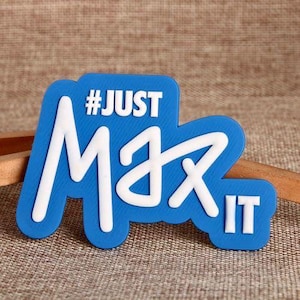 May include: A blue and white magnet with the text "#JUST Max IT". The magnet has a rounded rectangular shape with white lettering on a blue background. The text is in a bold, stylized font.