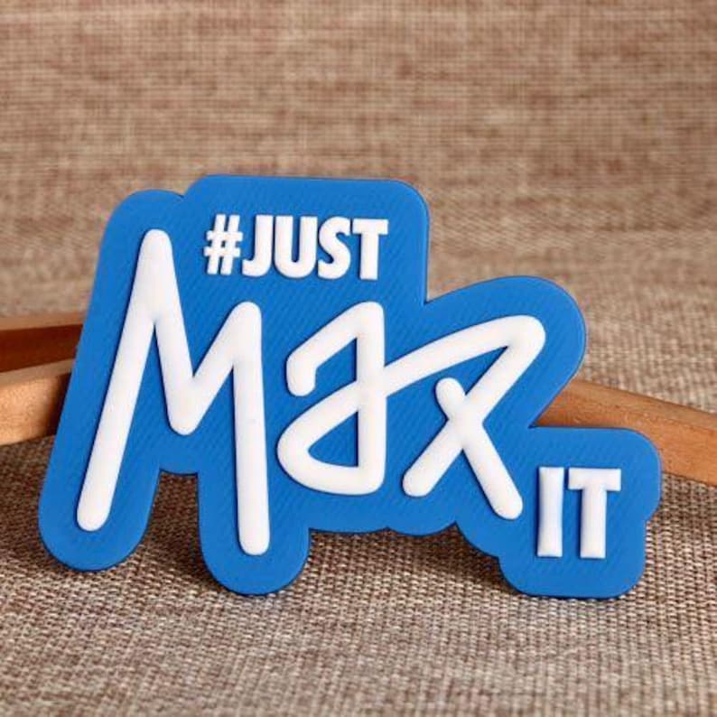 May include: A blue and white magnet with the text "#JUST Max IT". The magnet has a rounded rectangular shape with white lettering on a blue background. The text is in a bold, stylized font.