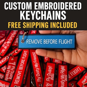Könnte beinhalten: Ein blaues, besticktes Schlüsselanhänger mit dem Text "REMOVE BEFORE FLIGHT" wird in einer Hand gehalten. Der Schlüsselanhänger hat einen silbernen Ring. Andere rote Schlüsselanhänger mit weißem Text befinden sich im Hintergrund. Der obere Teil des Bildes enthält den Text "CUSTOM EMBROIDERED KEYCHAINS".