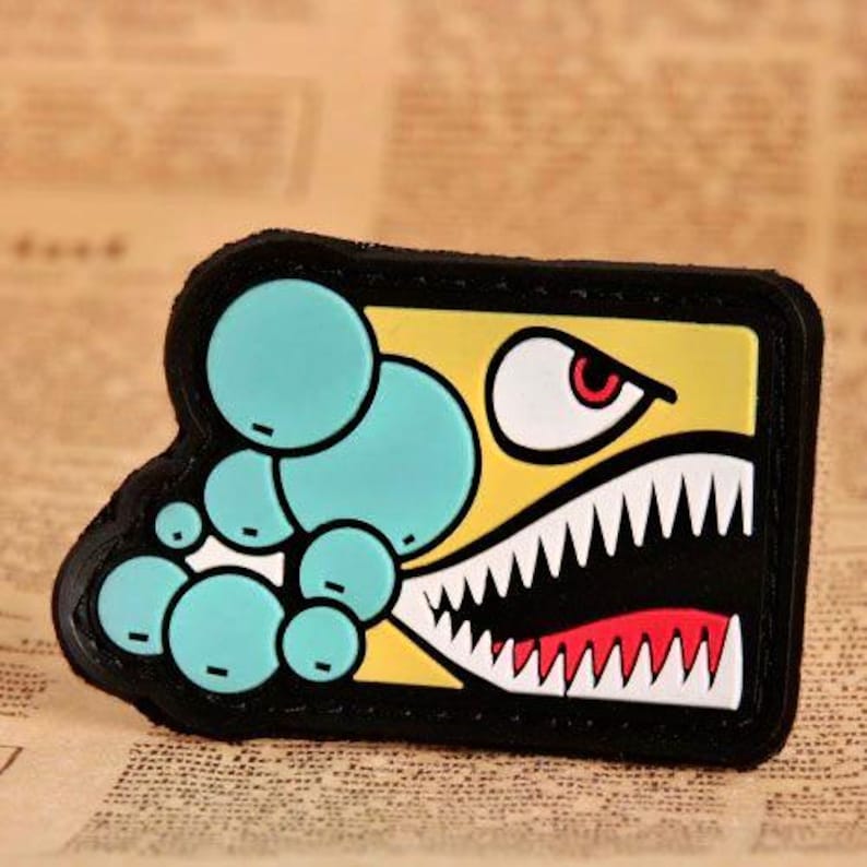 May include: A rectangular patch with a cartoon shark design. The patch features a yellow background with a shark's mouth, white teeth, and a red tongue. Teal bubbles are on the left side, and the patch is outlined in black.