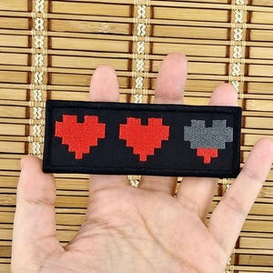 K&ouml;nnte beinhalten: Ein schwarzer, rechteckiger Aufn&auml;her mit drei pixeligen Herzen. Zwei Herzen sind rot, das dritte grau, was eine Gesundheitsanzeige darstellt. Der Aufn&auml;her ist ein Accessoire f&uuml;r Kleidung oder Taschen, f&uuml;r Gamer und Retro-Fans.