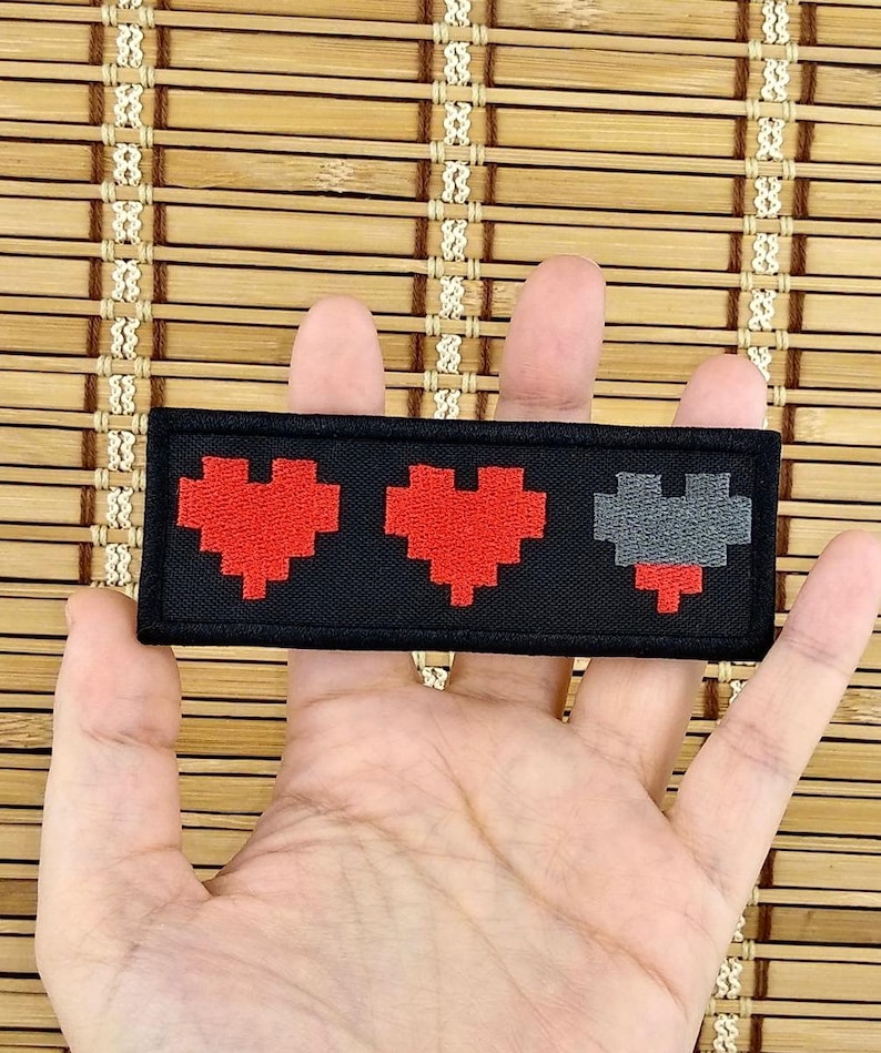 K&ouml;nnte beinhalten: Ein schwarzer, rechteckiger Aufn&auml;her mit drei pixeligen Herzen. Zwei Herzen sind rot, das dritte grau, was eine Gesundheitsanzeige darstellt. Der Aufn&auml;her ist ein Accessoire f&uuml;r Kleidung oder Taschen, f&uuml;r Gamer und Retro-Fans.