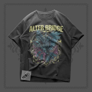May include: Dark gray t-shirt featuring the band name "ALTER BRIDGE" in gold lettering above a detailed graphic. The graphic depicts a stylized bird with blue and teal wings, set against a decorative background with purple and gold accents. The shirt has a crew neck.