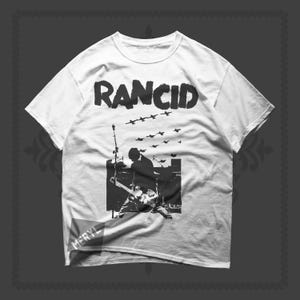 Puede incluir: Camiseta blanca con el nombre de la banda "RANCID" en letras negras y en negrita. El diseño muestra la silueta de un músico tocando la guitarra, con pájaros volando por encima. La camiseta tiene cuello redondo y mangas cortas.