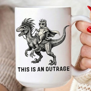以下が含まれることがあります： サングラスと王冠をつけた猫が恐竜に乗っている白のセラミックマグ。下には「THIS IS AN OUTRAGE」の文字がプリントされています。マグには白いハンドルが付いています。