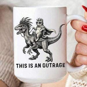 以下が含まれることがあります： サングラスと王冠をつけた猫が恐竜に乗っている白のセラミックマグ。イラストの下には「THIS IS AN OUTRAGE」の文字がプリントされています。