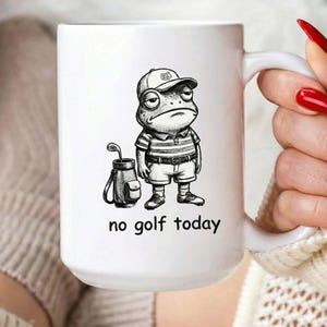 May include: White ceramic mug with a black and white illustration of a frog dressed in golf attire, with the text "no golf today". The frog is wearing a hat, striped shirt, shorts, and golf shoes. A golf bag and club are next to the frog.
