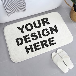 May include: A rectangular white bath mat with rounded corners featuring the black text YOUR DESIGN HERE printed in the center. The mat has a soft, plush, textured surface and is positioned on a gray tiled floor next to white slippers.