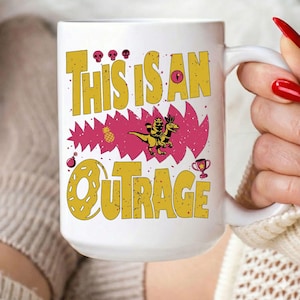 以下が含まれることがあります： 「THIS IS AN OUTRAGE」の文字が黄色で描かれた白いセラミックマグ。漫画の恐竜が馬に乗っている様子、パイナップル、その他のグラフィックが特徴です。マグには大きなハンドルが付いています。