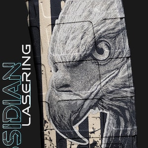 Pode incluir: Um carregador preto e bege com um design de bandeira americana desgastada e um gráfico detalhado da cabeça de uma águia. O texto "OBSIDIAN LASERING" está alinhado verticalmente no lado esquerdo.