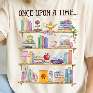 May include: A cream-colored t-shirt featuring a graphic print of four wooden shelves filled with colorful books and various fairy tale objects, including a rose in a glass dome, a golden lamp, a frog, a glass slipper, and the text ONCE UPON A TIME.