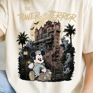 Può includere: Una t-shirt color crema con una grafica di Topolino vestito da fattorino che trasporta una valigia davanti all'Hollywood Tower Hotel. Il testo recita The Tower of Terror e The Hollywood Tower Hotel, con fantasmi e pipistrelli.