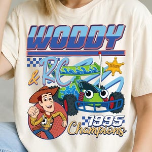 Può includere: Una t-shirt color crema con una stampa grafica di Woody e dell'auto RC. Il testo recita Woody & RC 1995 Champions. Il design include una stella da sceriffo gialla, motivi blu e verdi e un motivo a bandiera a scacchi sul davanti.