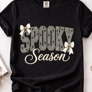 May include: A black short-sleeve t-shirt featuring the text SPOOKY in a grey spiderweb pattern with small black spiders, and the word Season in white cursive script. Two cream-colored bows are attached to the letters S and N.