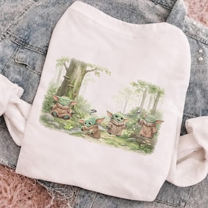 Op de afbeelding: Wit T-shirt met een grillige illustratie van vier groene buitenaardse wezens in een bos. De wezens dragen bruine gewaden en interageren. Het woord "cool" staat op de afbeelding.