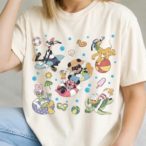 May include: Cream-colored t-shirt featuring a colorful graphic of Disney characters enjoying a summer day. The design includes Goofy, Mickey, Minnie, Pluto, Daisy, and Donald Duck with beach-themed elements like floats, sunglasses, and a beach ball.