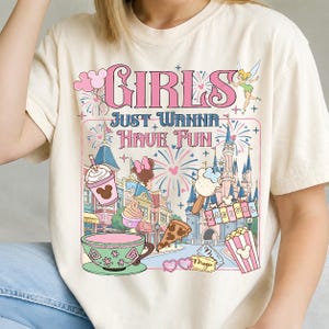 May include: Cream-colored t-shirt with the text "GIRLS JUST WANNA HAVE FUN" in pink. The graphic includes a cartoon castle, fireworks, and various food items like ice cream and popcorn. A fairy is also featured.