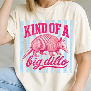 May include: Cream-colored t-shirt with a pink armadillo graphic and the text "KIND OF A big dillo" in pink lettering. The armadillo is centered on a light blue striped background.