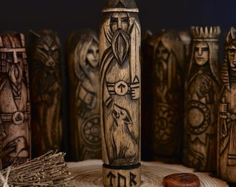 May include: Wooden carved figures of Norse gods and symbols. The central figure depicts a bearded man with a wolf, holding a sword and arrow. Other figures include women and animals. The figures are on a wooden slice with runes.