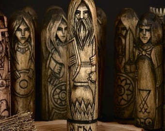 May include: A collection of intricately carved wooden figures, each depicting a stylized human form with detailed facial features and clothing. The central figure, with the word "AGPA" at its base, stands on a wooden slice with several round, dark brown tokens.