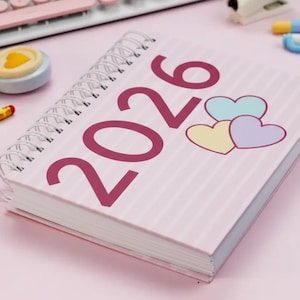 Może przedstawiać: Różowy spiralny planer na rok 2026 z bordowymi literami. Okładka zawiera trzy pastelowe serca i pionowe paski. Planer jest otwarty, ukazując białe strony.