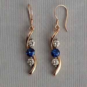 May include: Rose gold-tone earrings with a curved design. Each earring features a sparkling blue gemstone, flanked by two clear, round gemstones. The earrings have a hook closure and are displayed against a gray background.