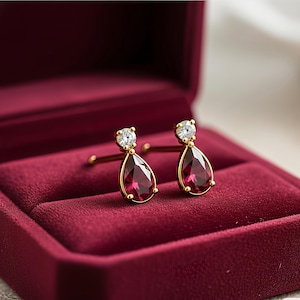May include: A pair of gold-colored earrings with a teardrop-shaped, deep red gemstone and a small, round, clear stone at the top. The earrings are displayed in a burgundy velvet jewelry box.
