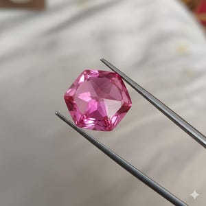 Peut inclure: Une pierre précieuse rose hexagonale à facettes est maintenue par une pince argentée. La gemme a de multiples facettes qui reflètent la lumière, créant un effet scintillant. La pierre est d'un rose vif.