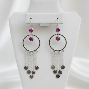 Peut inclure: Une paire de boucles d'oreilles pendantes argentées. Chaque boucle d'oreille comporte un grand cercle, une petite chaîne avec des breloques circulaires et deux cristaux magenta. Les boucles d'oreilles sont exposées sur un présentoir à bijoux blanc.