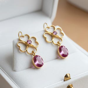 Peut inclure: Une paire de boucles d'oreilles dorées avec un motif floral. Chaque boucle d'oreille présente un dessus en forme de trèfle avec une petite pierre rose et une plus grande pierre rose ovale pendante. Les boucles d'oreilles sont présentées dans un écrin blanc.