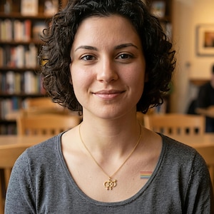 May include: A gold-toned necklace featuring a pendant with two interlocking female gender symbols. The necklace is worn over a dark gray scoop-neck shirt. A small, horizontal rainbow-colored stripe graphic is visible on the upper chest area.