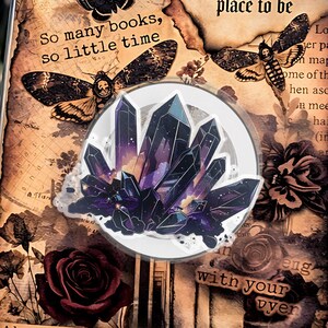 May include: Decorative book with a vintage aesthetic. The cover features a crystal design, moth illustrations, and text that reads "So many books, so little time" and "Between the pages of a book is a lovely place to be." A key and skull charm hang from the book.