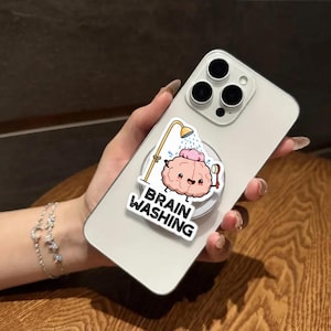 May include: A white smartphone with a pop-up grip featuring a cartoon brain under a shower, with the words "BRAIN WASHING" in white. The brain is pink and smiling. The phone is held by a person.