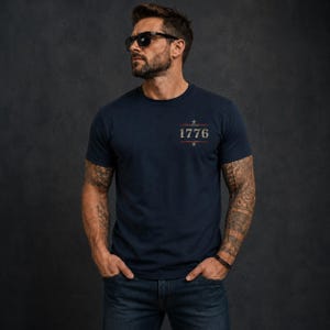May include: A navy blue short-sleeved t-shirt featuring a graphic on the chest with the text 1776 centered between two horizontal lines and two small stars. The design has a distressed, weathered appearance in muted red, white, and blue tones.