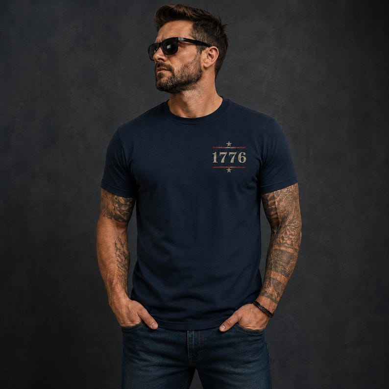 May include: A navy blue short-sleeved t-shirt featuring a graphic on the chest with the text 1776 centered between two horizontal lines and two small stars. The design has a distressed, weathered appearance in muted red, white, and blue tones.