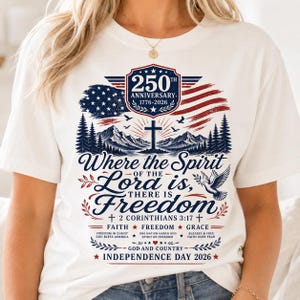 May include: A white t-shirt featuring a patriotic graphic with a red, white, and blue American flag, a mountain landscape, a cross, and a dove. The text reads 250 Anniversary 1776-2026, Where the Spirit of the Lord is, there is Freedom, 2 Corinthians 3:17.