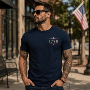 May include: A navy blue short-sleeved t-shirt featuring the text 1776 printed in a light, distressed font on the chest. The design includes a small star above and below the numbers, flanked by horizontal lines, creating a centered graphic.