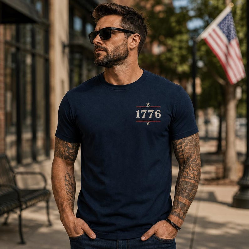 May include: A navy blue short-sleeved t-shirt featuring the text 1776 printed in a light, distressed font on the chest. The design includes a small star above and below the numbers, flanked by horizontal lines, creating a centered graphic.