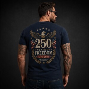 May include: A navy blue t-shirt featuring a graphic on the back with a bald eagle, two American flags, and the text 250 YEARS OF FREEDOM 1776-2026 UNITED STATES OF AMERICA, all framed by a gold laurel wreath and a series of small stars.