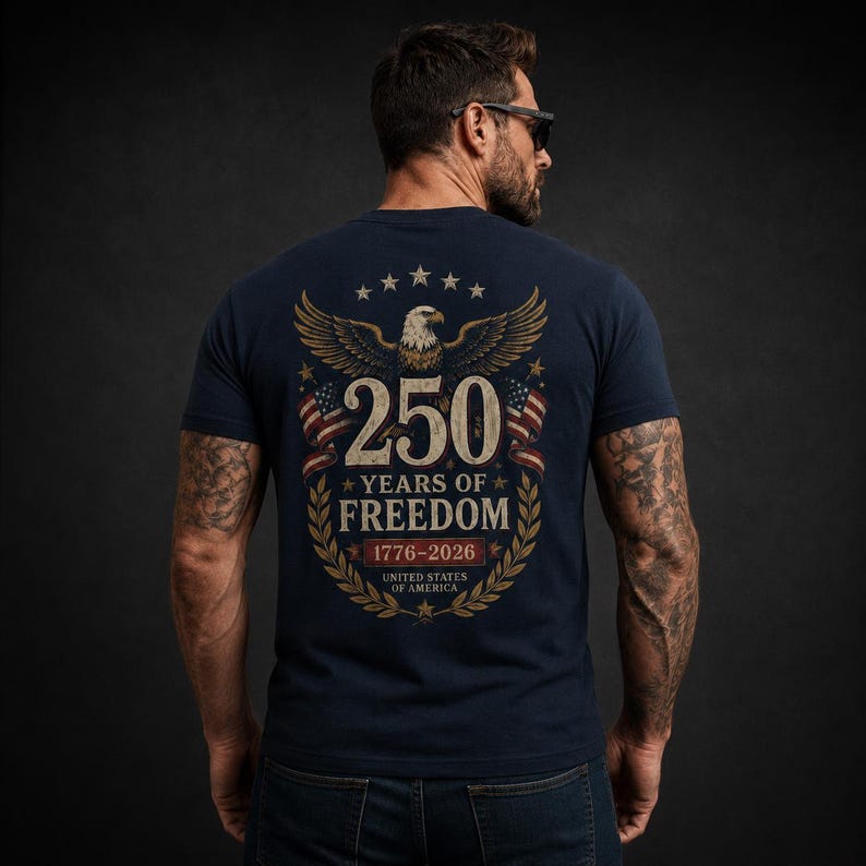 May include: A navy blue t-shirt featuring a graphic on the back with a bald eagle, two American flags, and the text 250 YEARS OF FREEDOM 1776-2026 UNITED STATES OF AMERICA, all framed by a gold laurel wreath and a series of small stars.