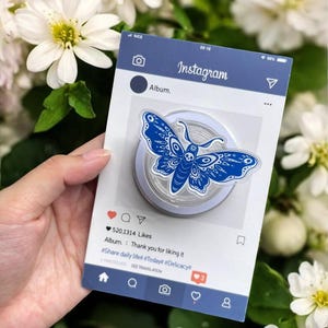 May include: A blue and white butterfly-shaped phone grip is displayed on a card designed to look like an Instagram post. The card has the Instagram logo and the text "Album. Thank you for liking it". The background is blurred with white flowers.